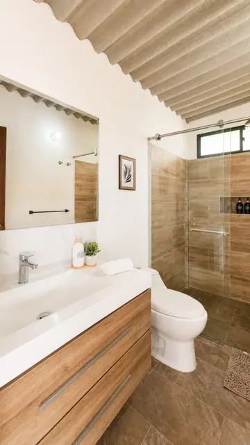 Baño con lavamanos sobre mesón blanco, mueble en madera y ducha con vidrio